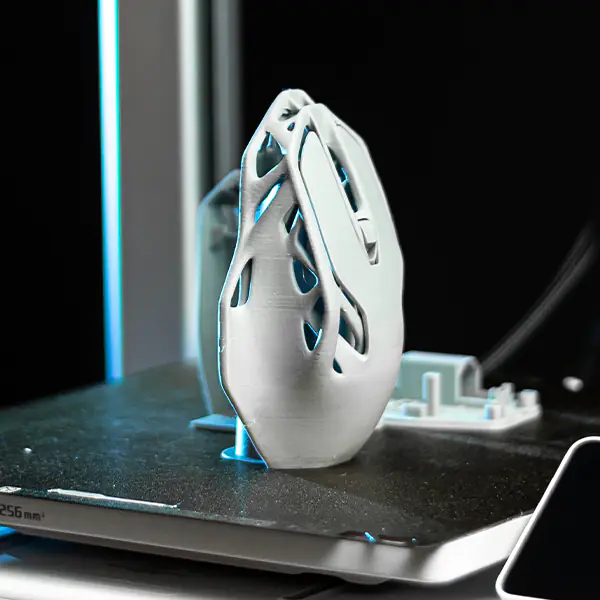 Nahaufnahme eines 3D-gedruckten Prototyps einer ergonomischen Computermaus mit offener Wabenstruktur, der auf der Bauplatte eines 3D-Druckers steht.