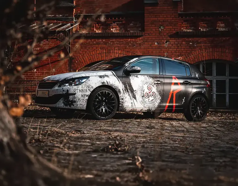 Keltische Vollfolierung an einem Peugeot 308 von foxdot.me mit Runen, Triskele-Symbol und roten Akzenten vor Backsteinkulisse.